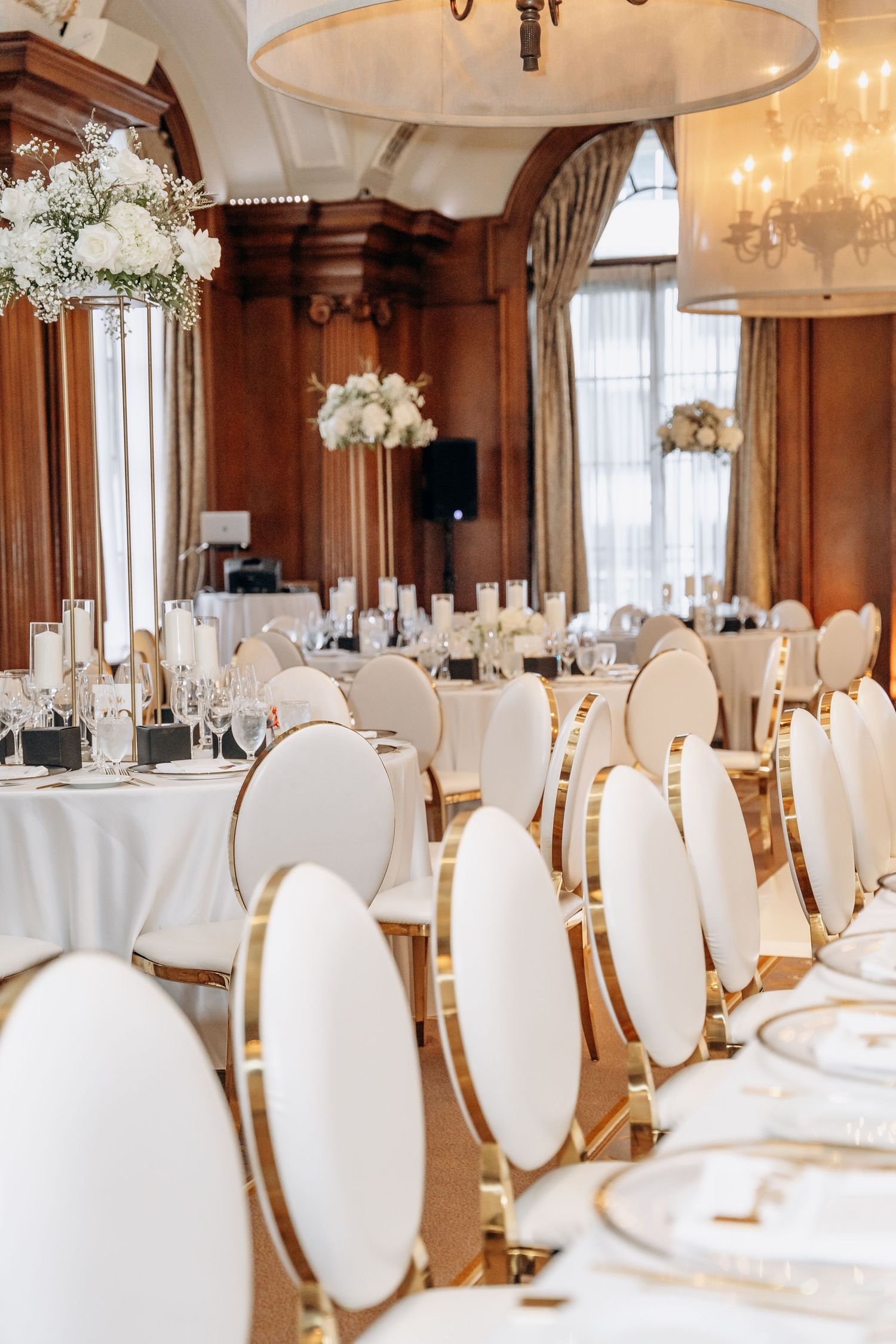 Elegant wedding reception setup with white floral centerpieces, gold chairs and white linens in a wood-paneled ballroom.