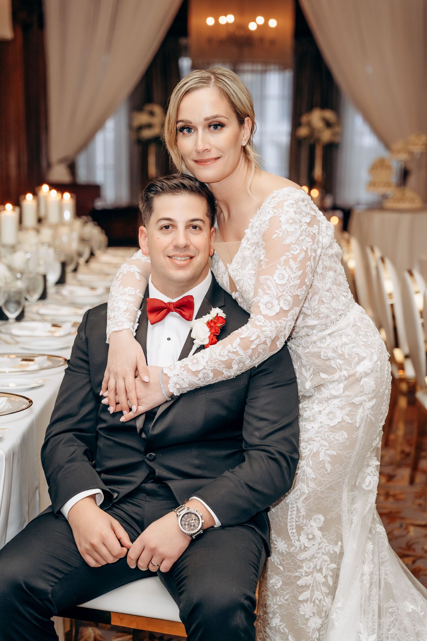 A couple in wedding attire sits together at an elegant reception with candlelit tables in the background.