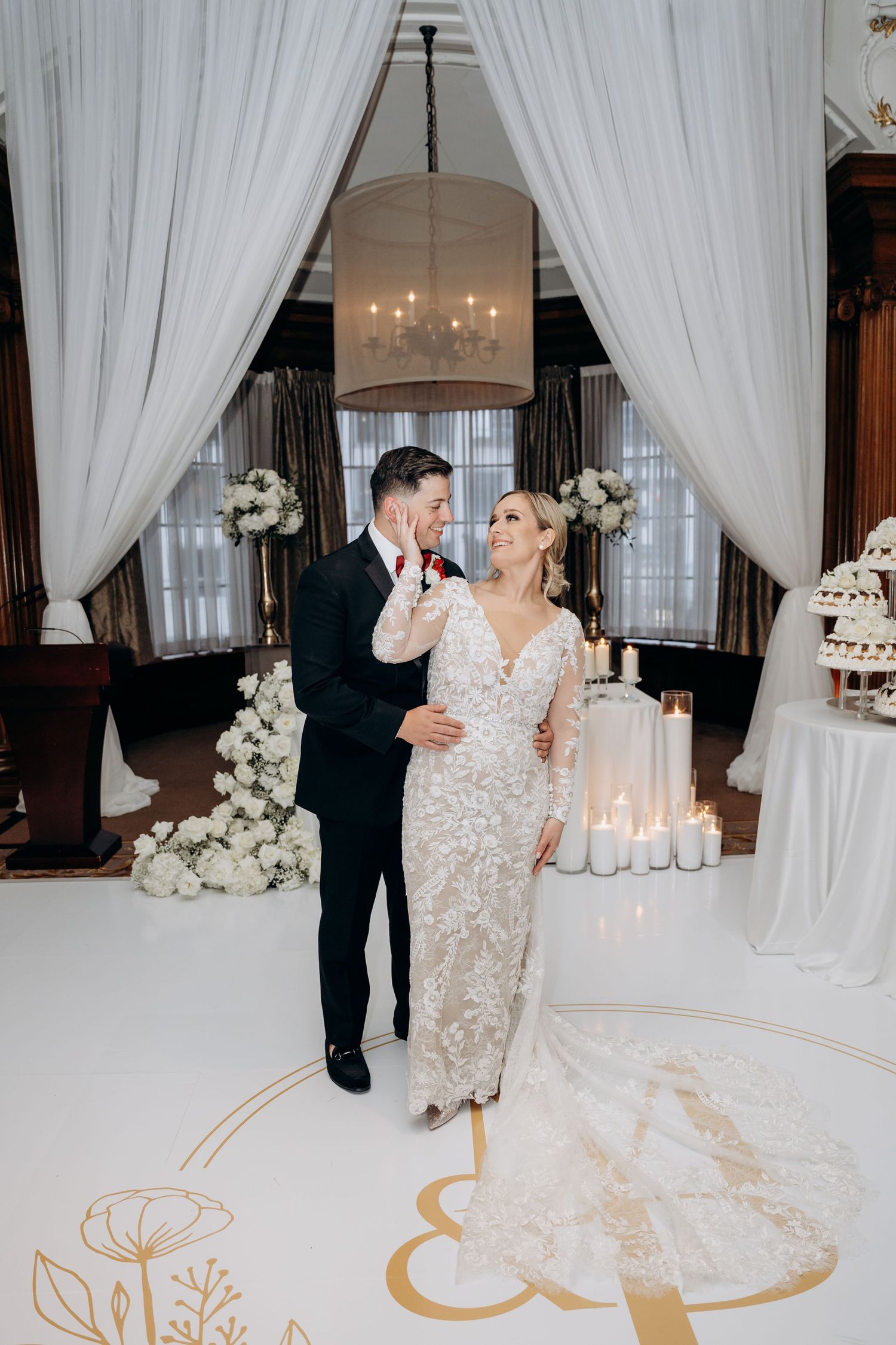 Elegant wedding reception venue with white drapes, floral arrangements, and a custom gold monogram design on the dance floor.