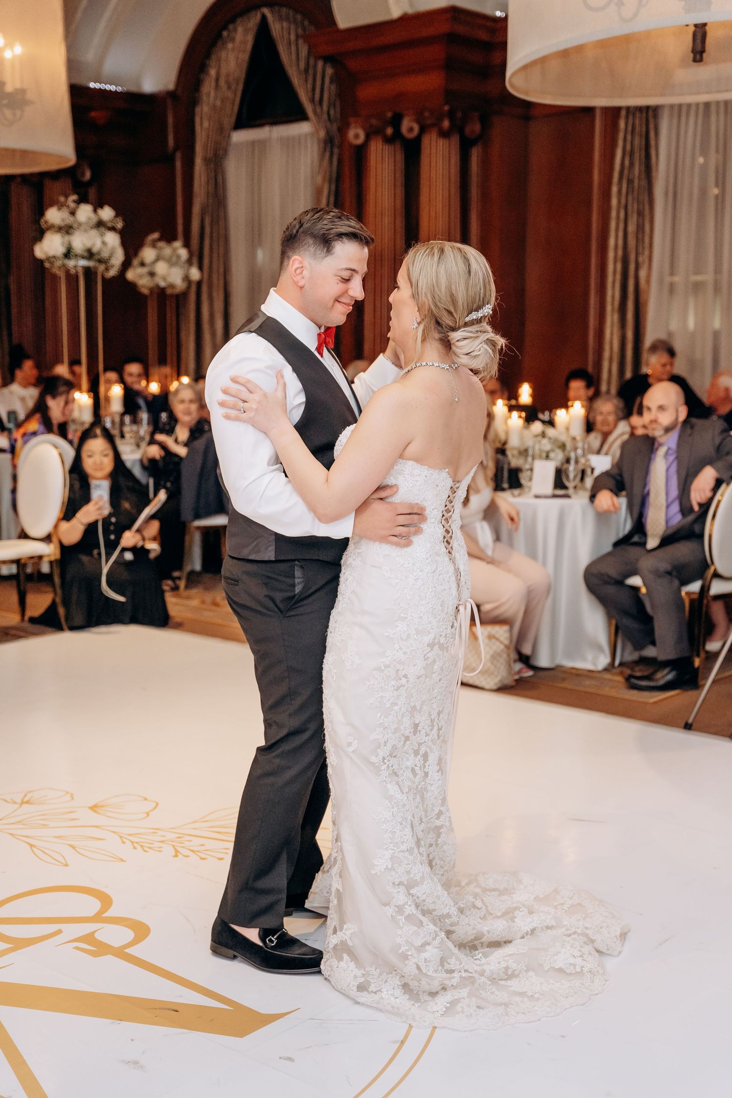 Couple shares their first dance at an elegant wedding reception with white decor and guests watching from tables.