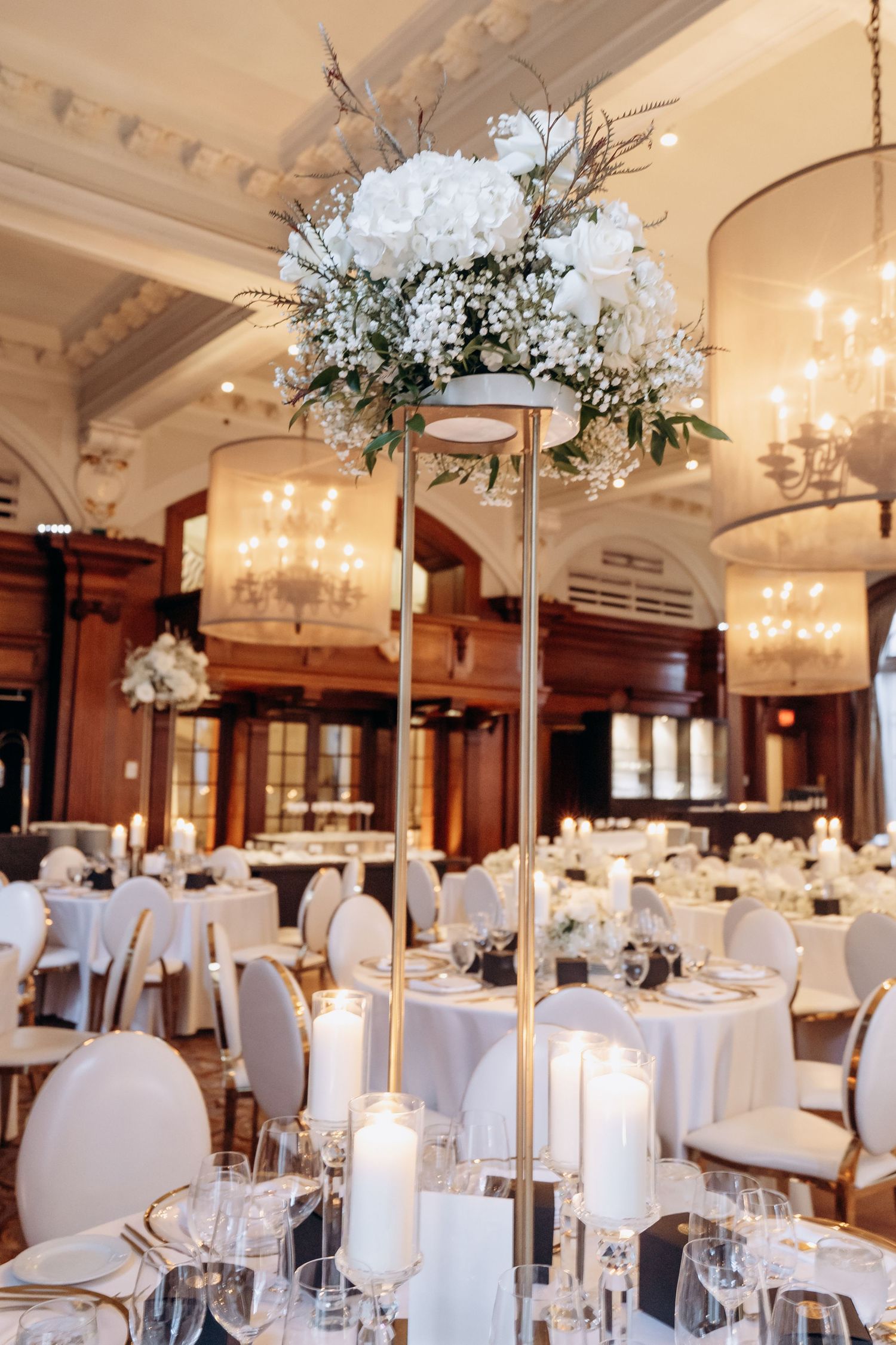 Elegant wedding reception ballroom with white chairs, tall floral centerpieces, and crystal chandeliers creating a luxurious atmosphere.