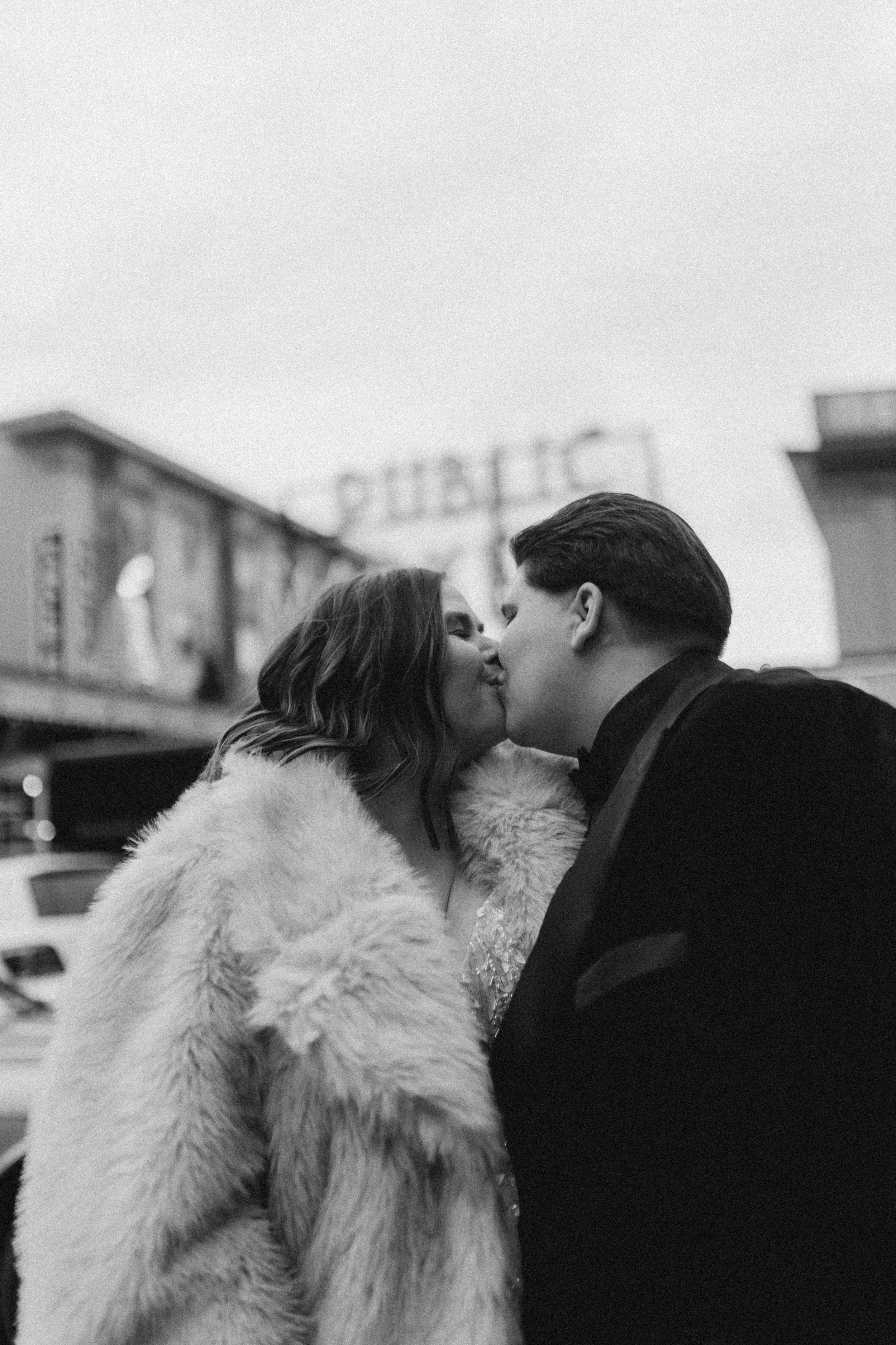 A couple sharing a romantic kiss in winter, with one wearing a white fur coat, shown in black and white photography.