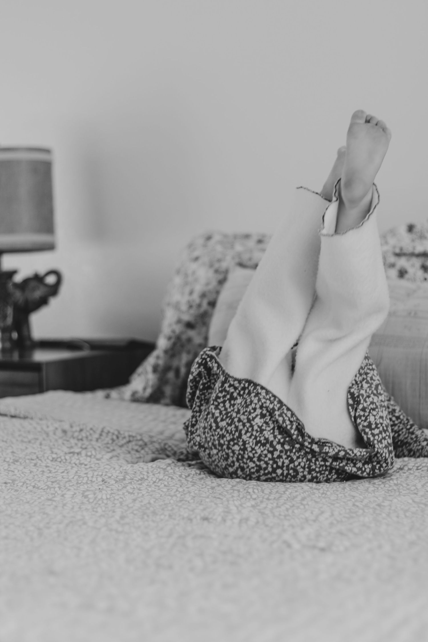 Black and white photo of feet raised up on a cozy bed with floral pillows and blankets in a bedroom | Kids Birthday Photo Sessions