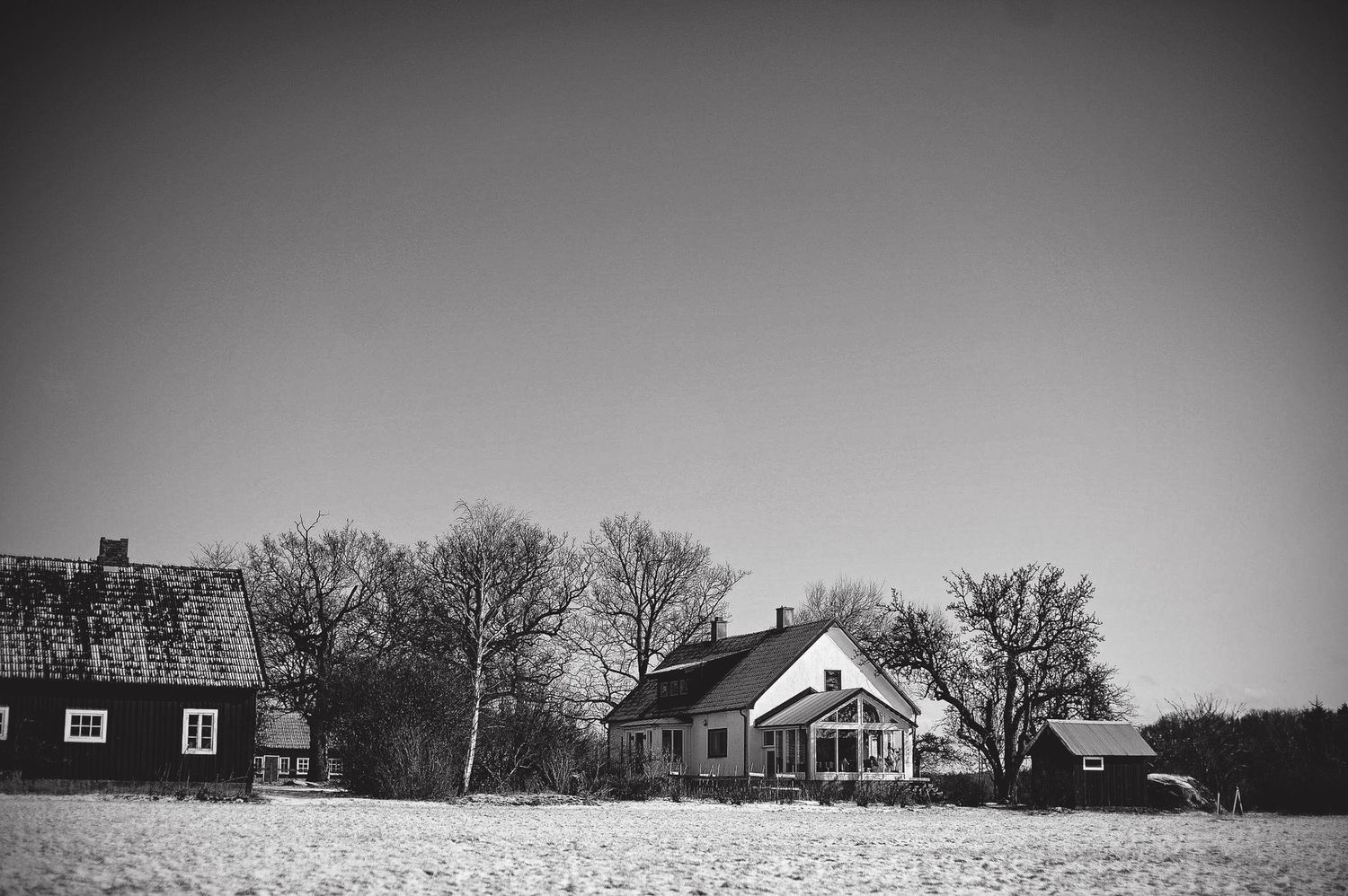A black and white landscape photograph of two rural houses in a snowy field with bare trees in the background.