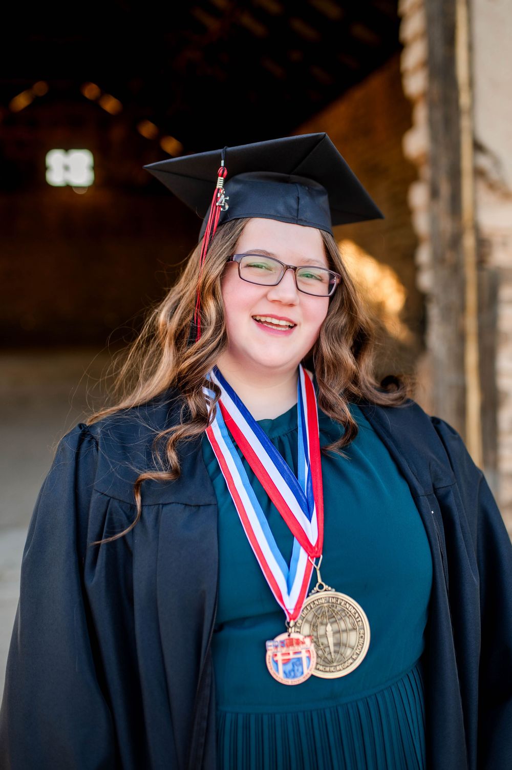 Proud graduate in cap and gown wearing honor medals smiles for senior portrait in warm light.