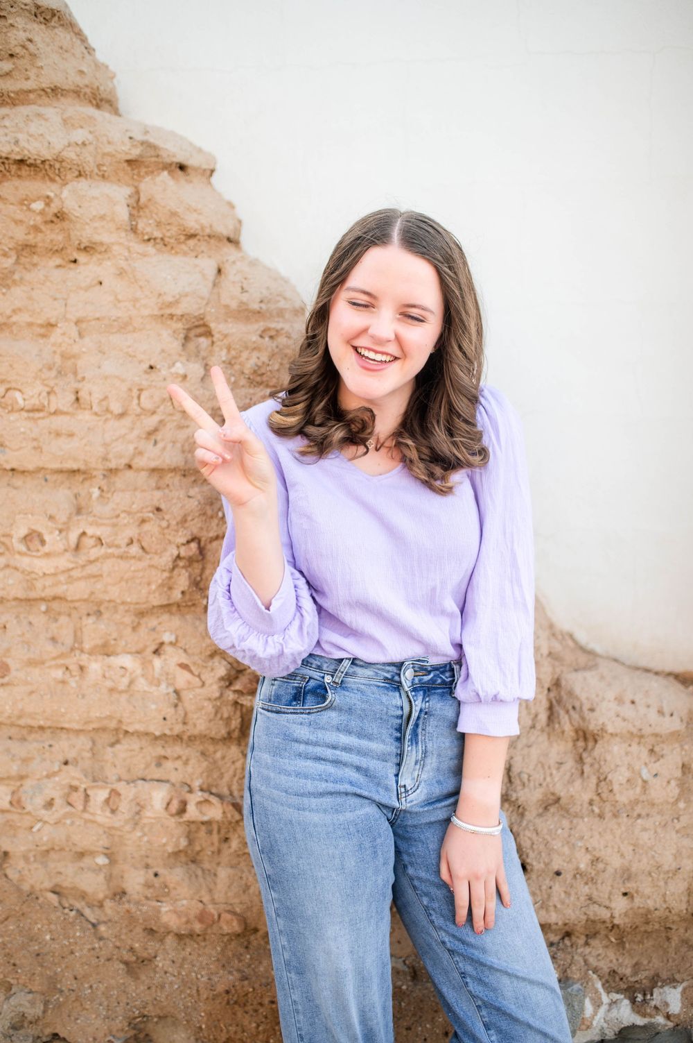 Cheerful teenage girl in lavender top and jeans flashes peace sign against adobe wall backdrop.