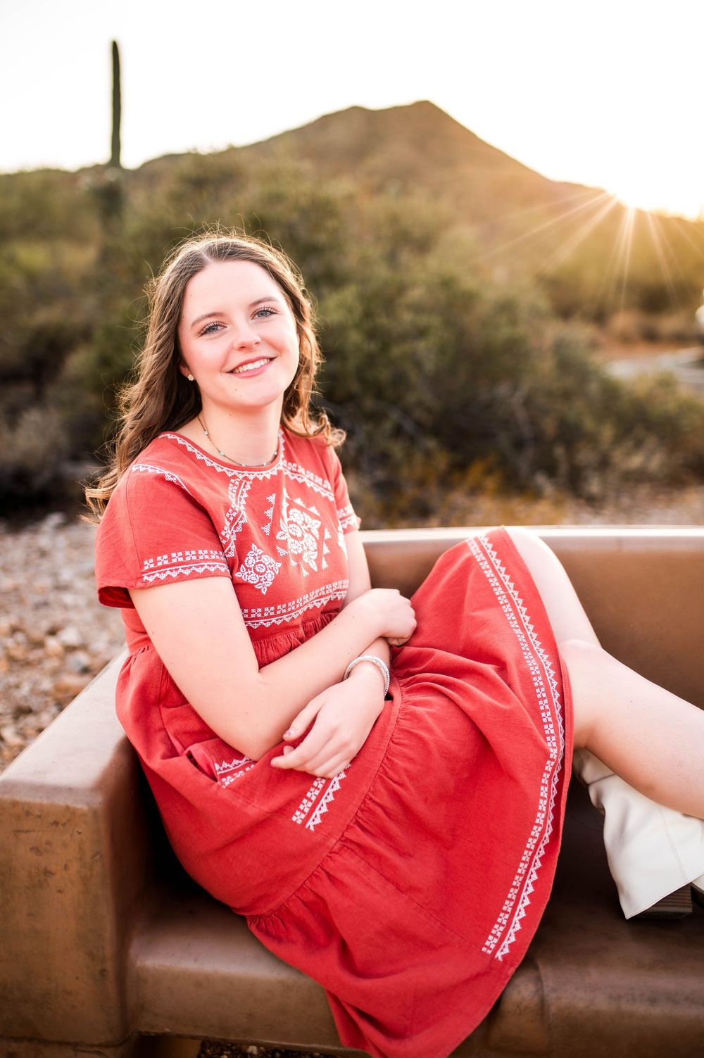 Smiling young woman in red embroidered dress sits on leather bench with Arizona desert sunset behind her.