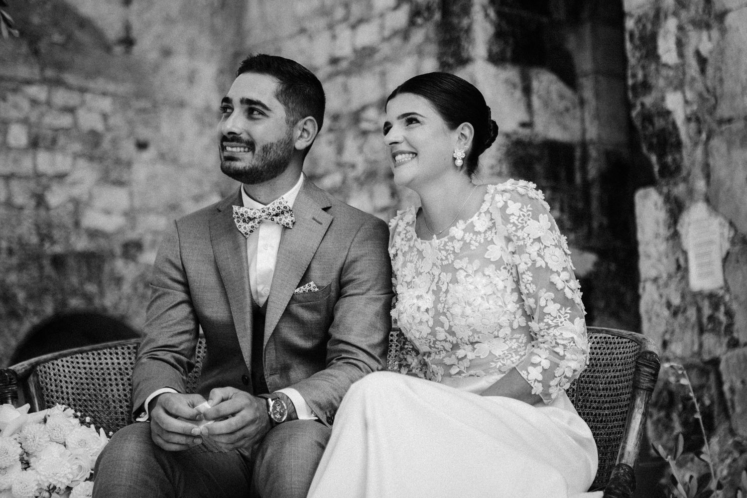 Bride and groom seated together during their wedding ceremony, smiling joyfully in a historic stone setting.