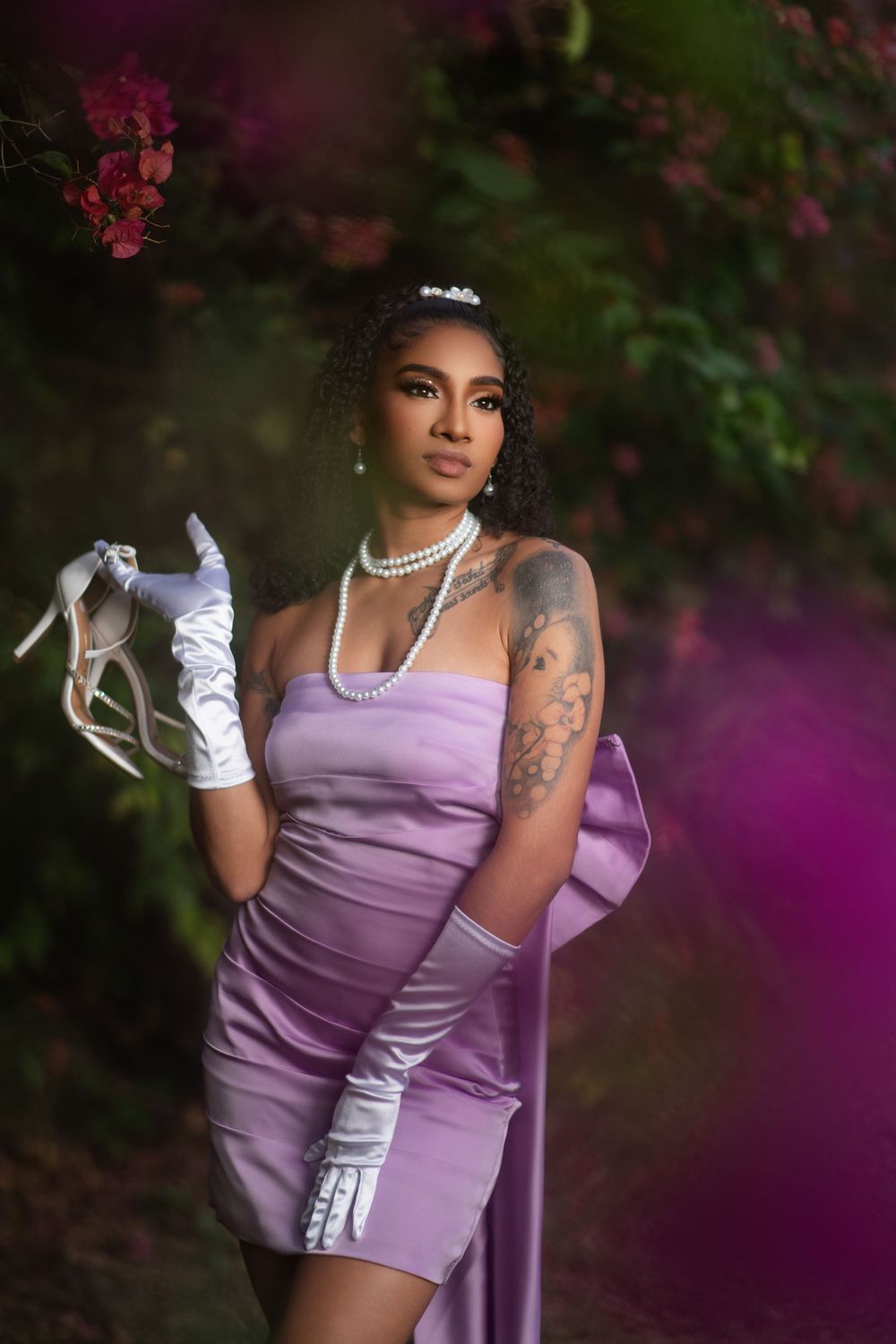 A person poses in a lavender dress and white gloves against a backdrop of vibrant pink and purple flowers in a garden setting.