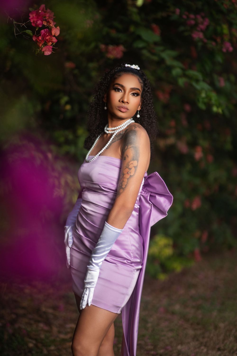 Fashion portrait in a lilac dress with long white gloves against a backdrop of pink flowers and greenery.