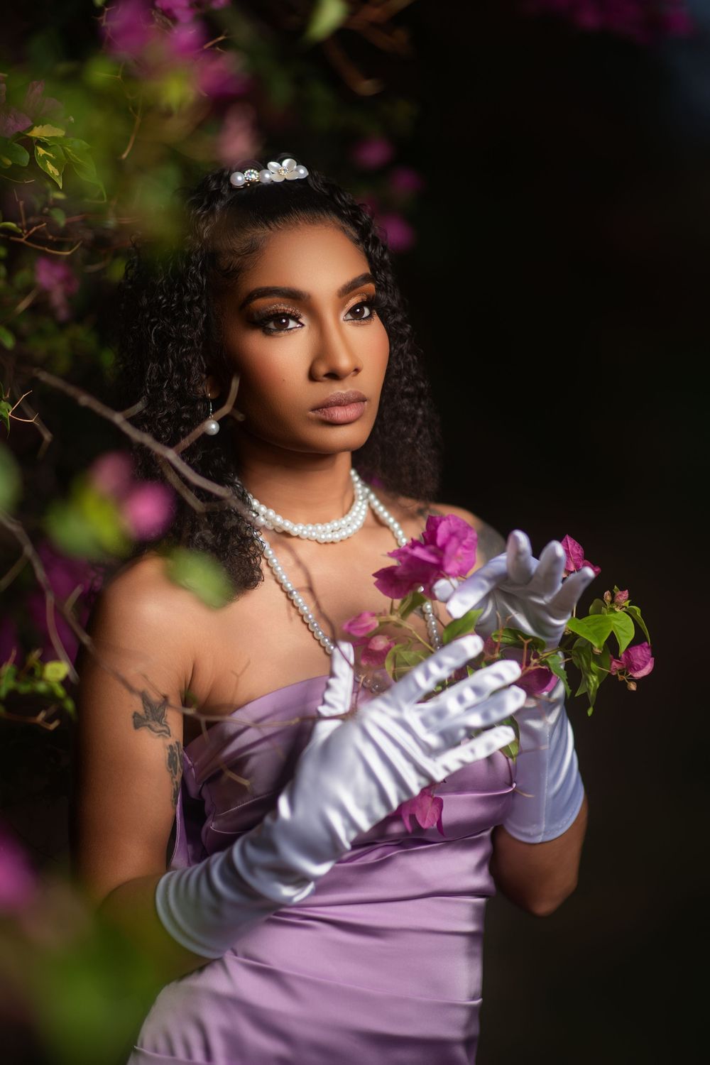 A portrait session in a garden features someone wearing a purple gown and white gloves among blooming flowers.