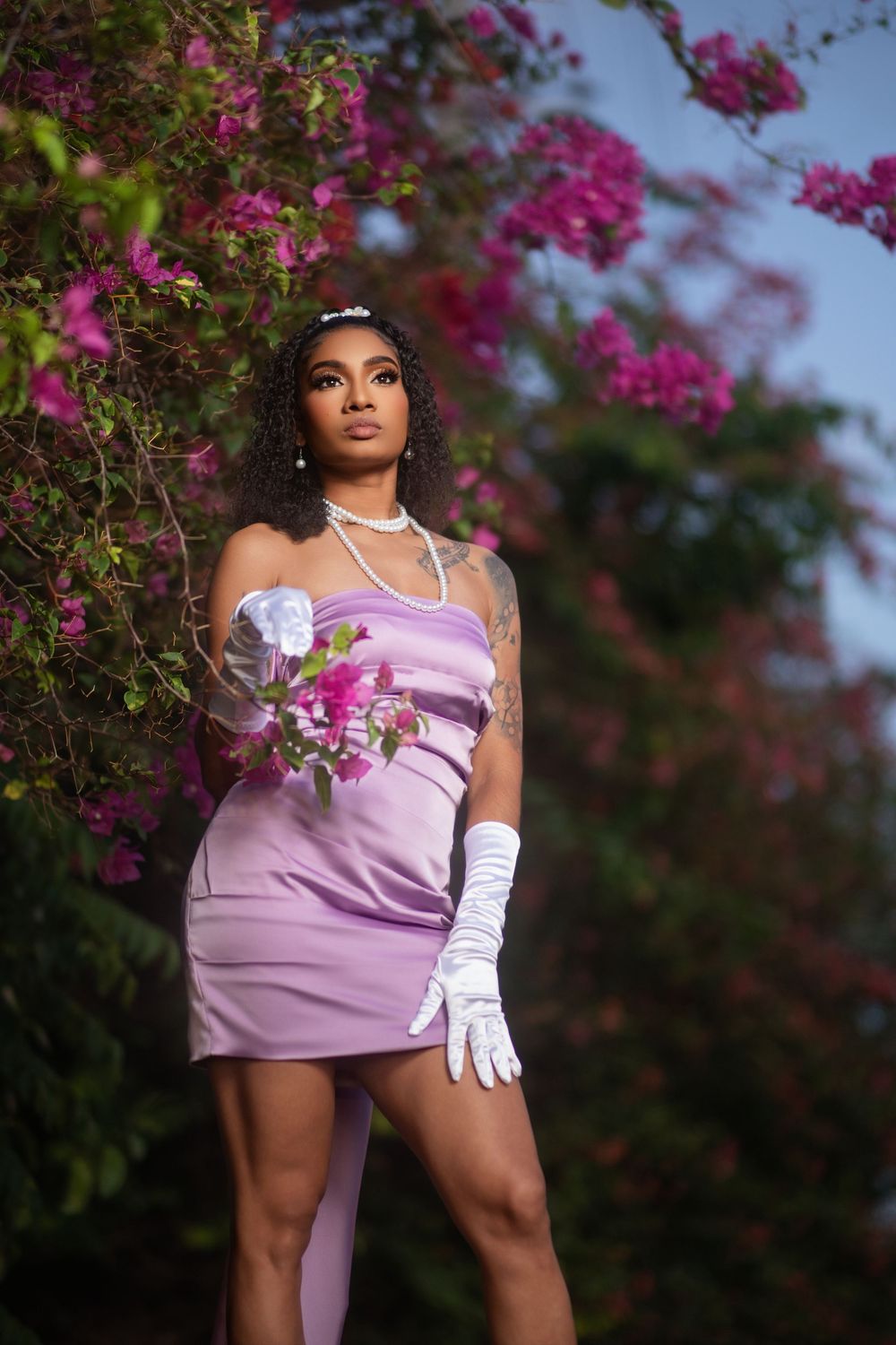 Fashion portrait of someone in a lavender dress and white gloves posing against vibrant pink bougainvillea flowers.