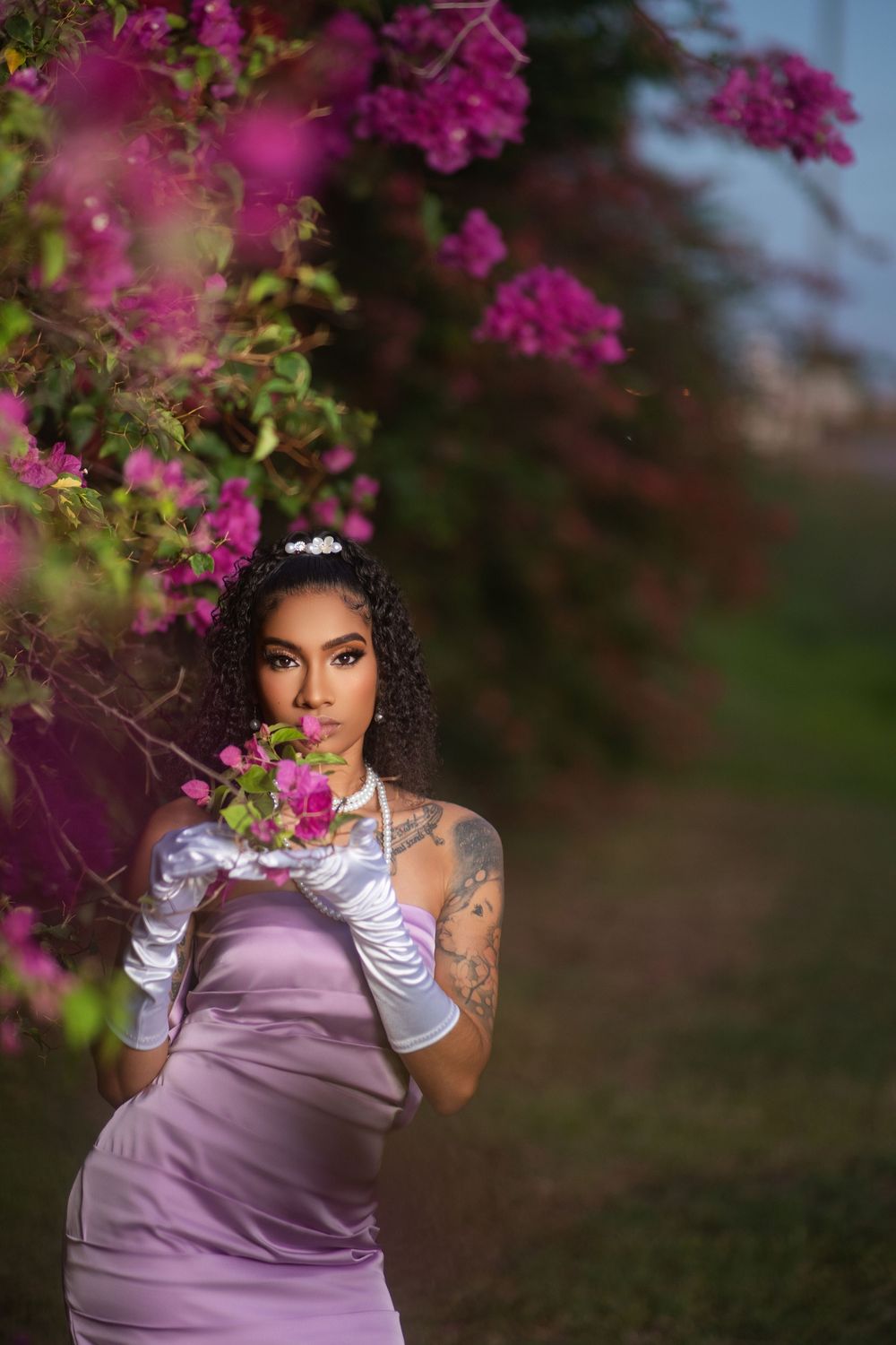 Elegant photoshoot featuring white gloves and lavender dress against blooming magenta flowers at dusk.