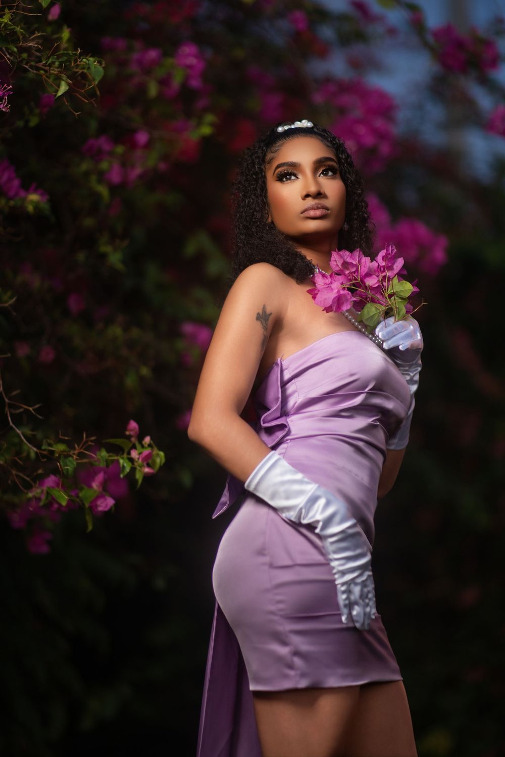 A maternity photoshoot in a garden features a purple satin dress against vibrant magenta bougainvillea flowers.