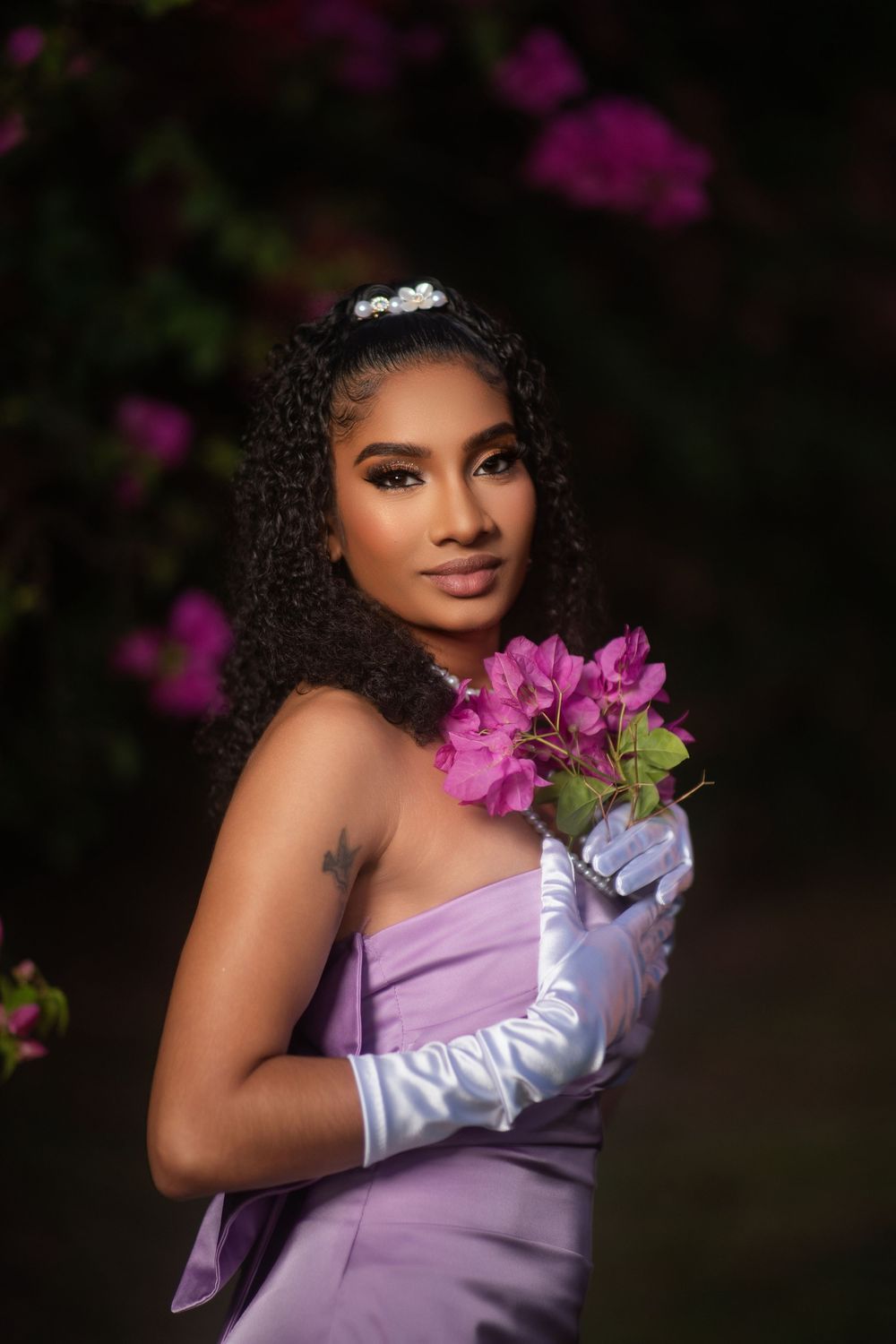 A figure in a lavender dress and white gloves poses elegantly with purple flowers against a dark background.