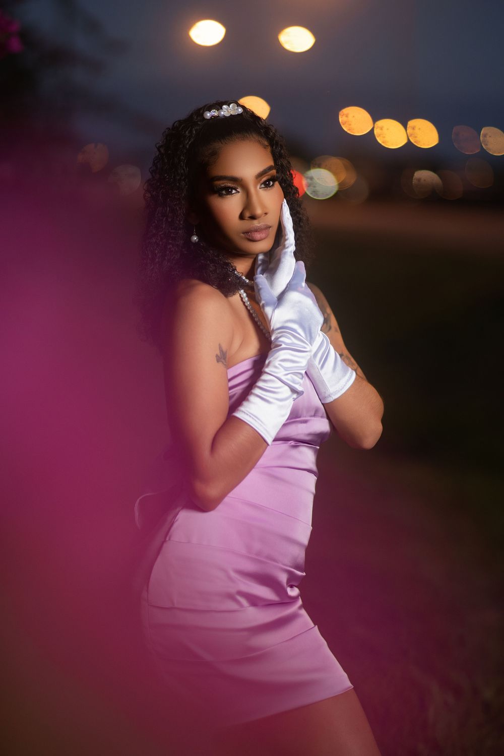 Evening photoshoot with bokeh lights creating dreamy atmosphere around figure in lavender dress.