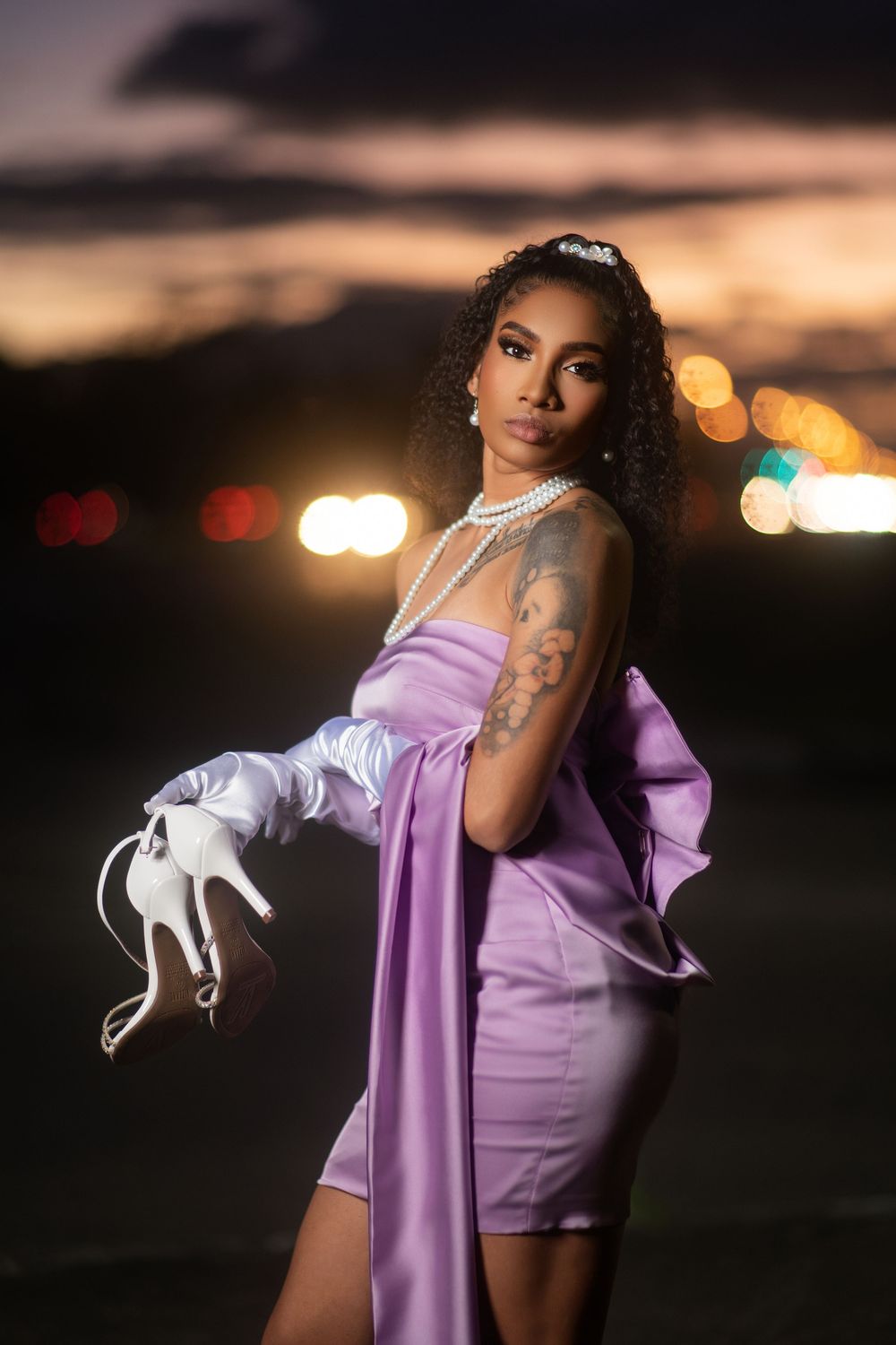 A figure in a lavender dress poses dramatically against a dusky sky with bokeh lights in the background.
