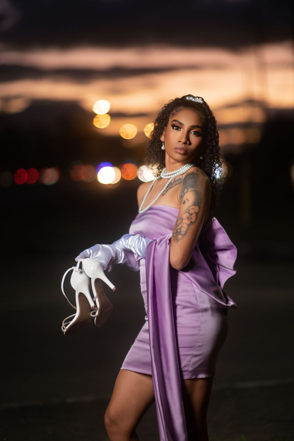 A figure in a purple dress poses dramatically against a dark evening sky with bokeh lights in the background.