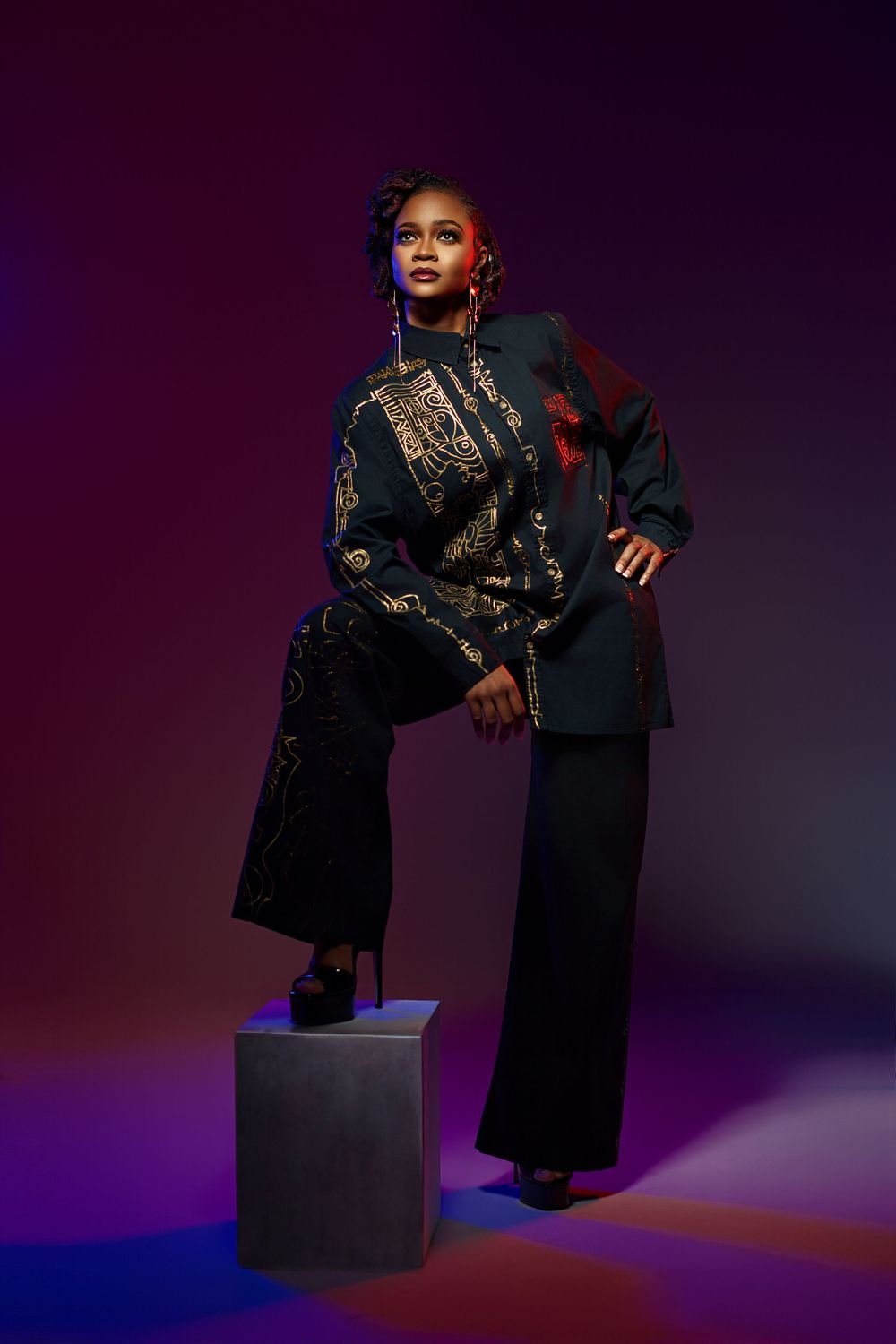 Fashion model in black lace dress and boots poses dramatically on cube against dark purple studio backdrop.