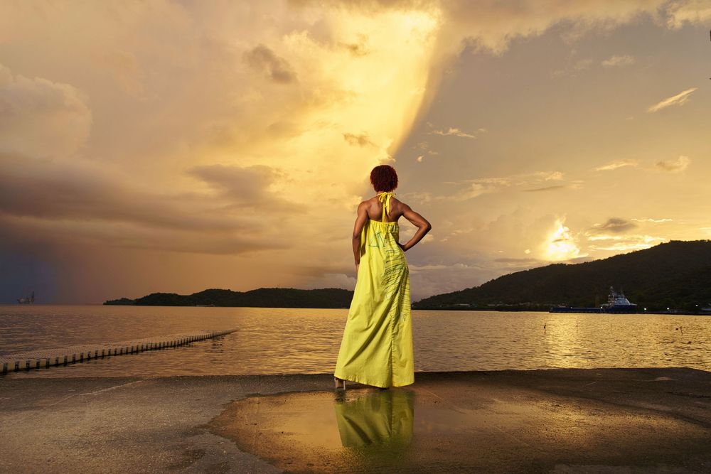 Silhouette in flowing lime dress poses against golden sunset reflecting on calm ocean waters.