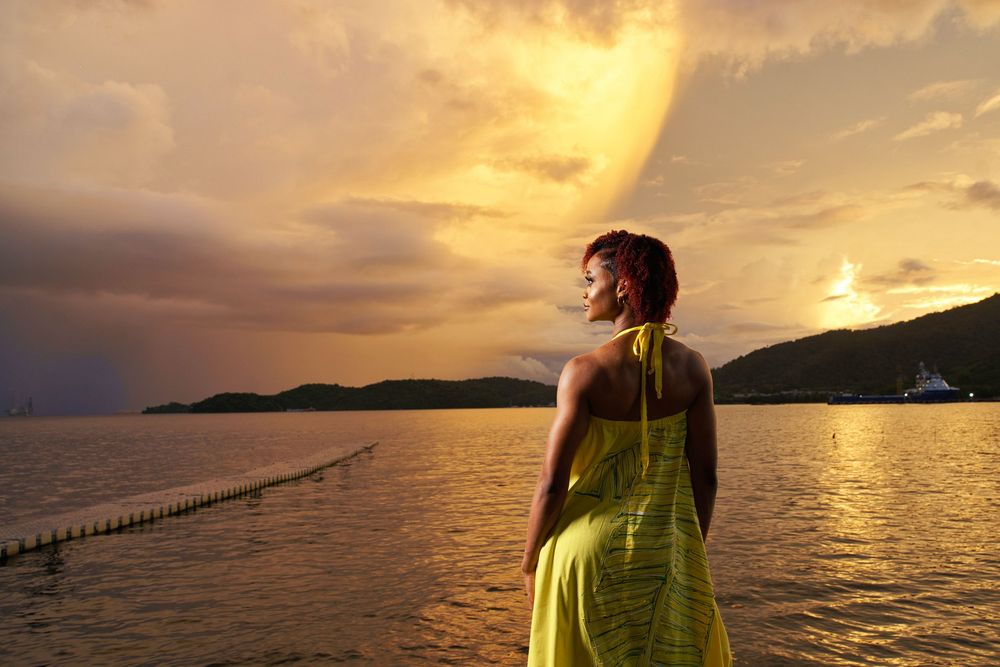 Figure in a bright yellow dress stands at waters edge during a dramatic golden sunset over mountains and ocean.