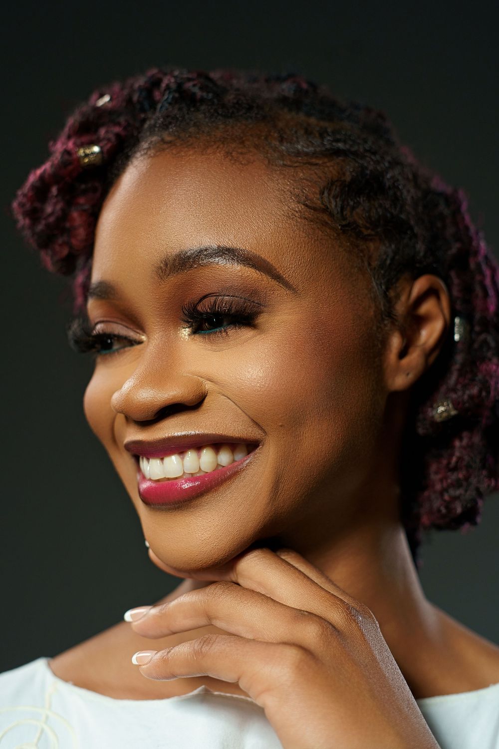 Professional beauty portrait showing bright smile and burgundy curls against dark background.