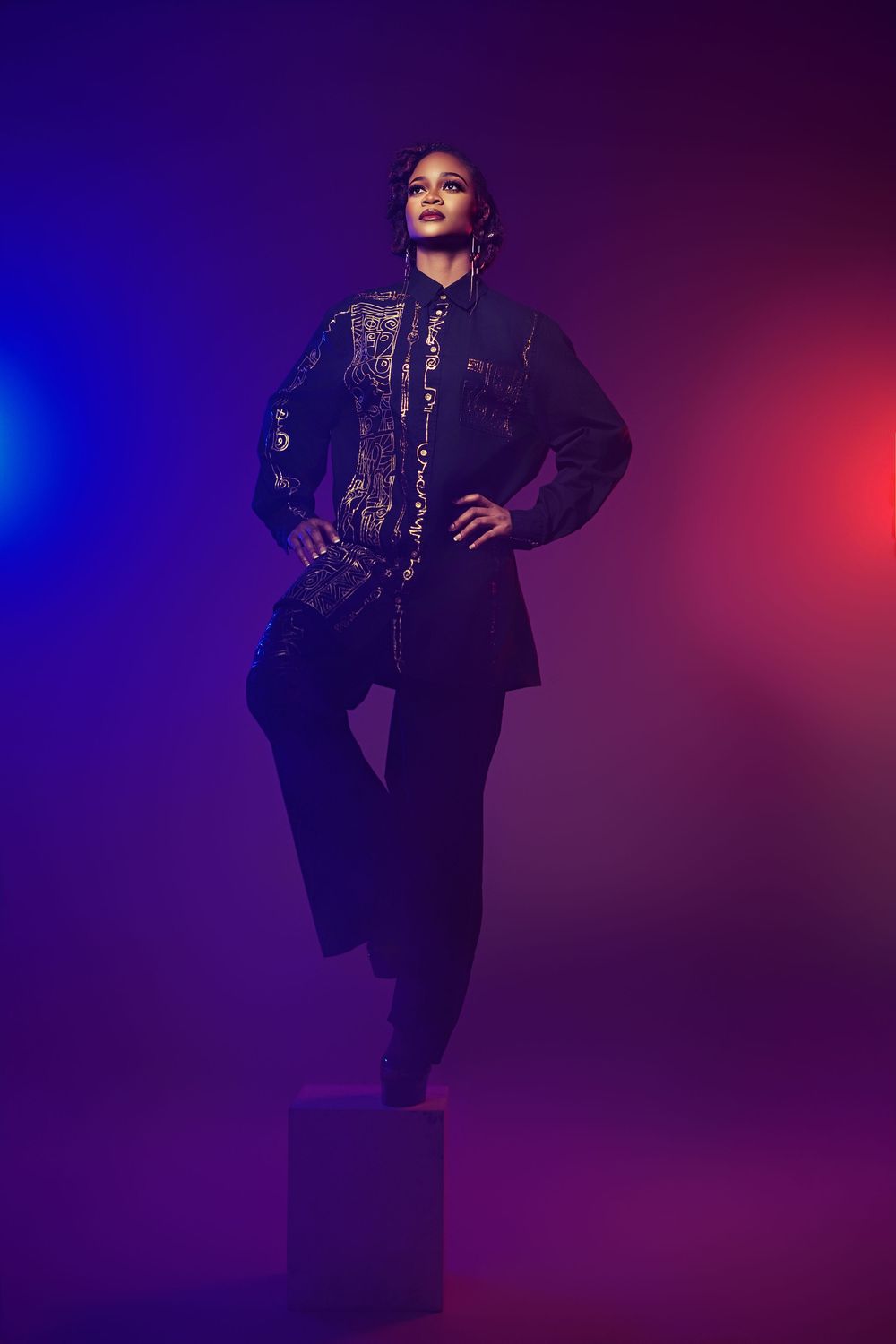 Figure in a dark sequined blazer and pants poses dramatically against vibrant purple and red lighting.
