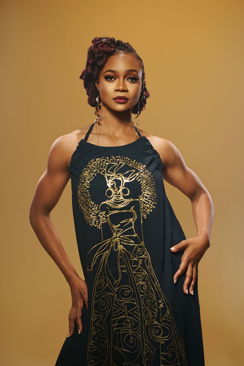 Fashion model poses in elegant black dress with gold line art design against warm brown backdrop.