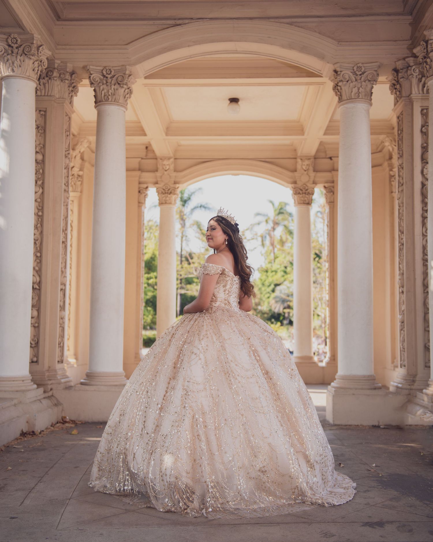 A bride in a flowing lace ballgown poses elegantly under a classical colonnade with ornate architectural details.