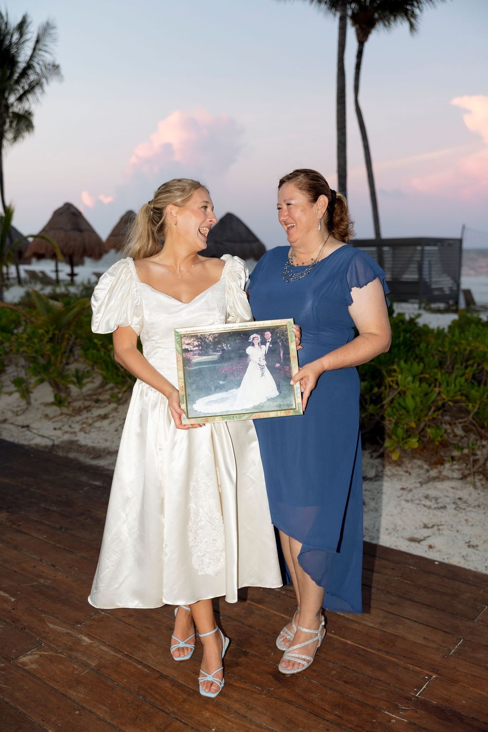Two people on a wooden deck hold a framed photo during sunset at a beach resort.
