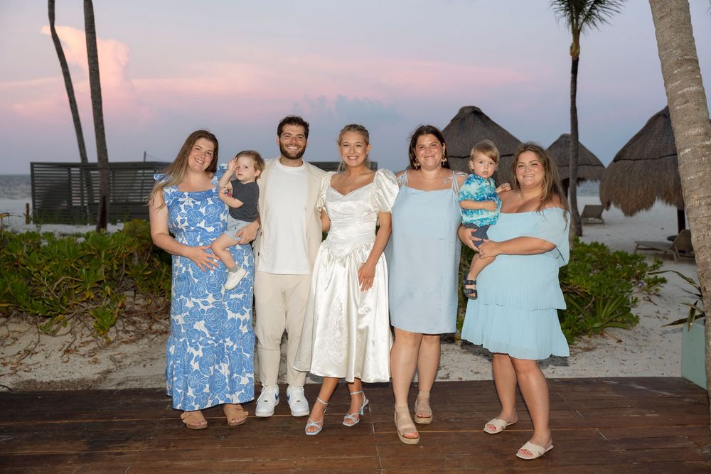A group in coordinated blue and white outfits poses together on a beach boardwalk at sunset with palm trees and thatched huts.