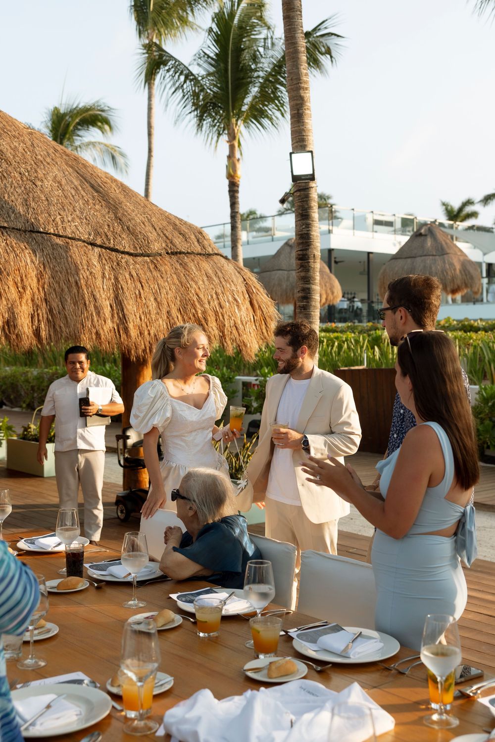 Guests enjoying a casual outdoor dining experience at a tropical restaurant with thatched roof palapas and palm trees.