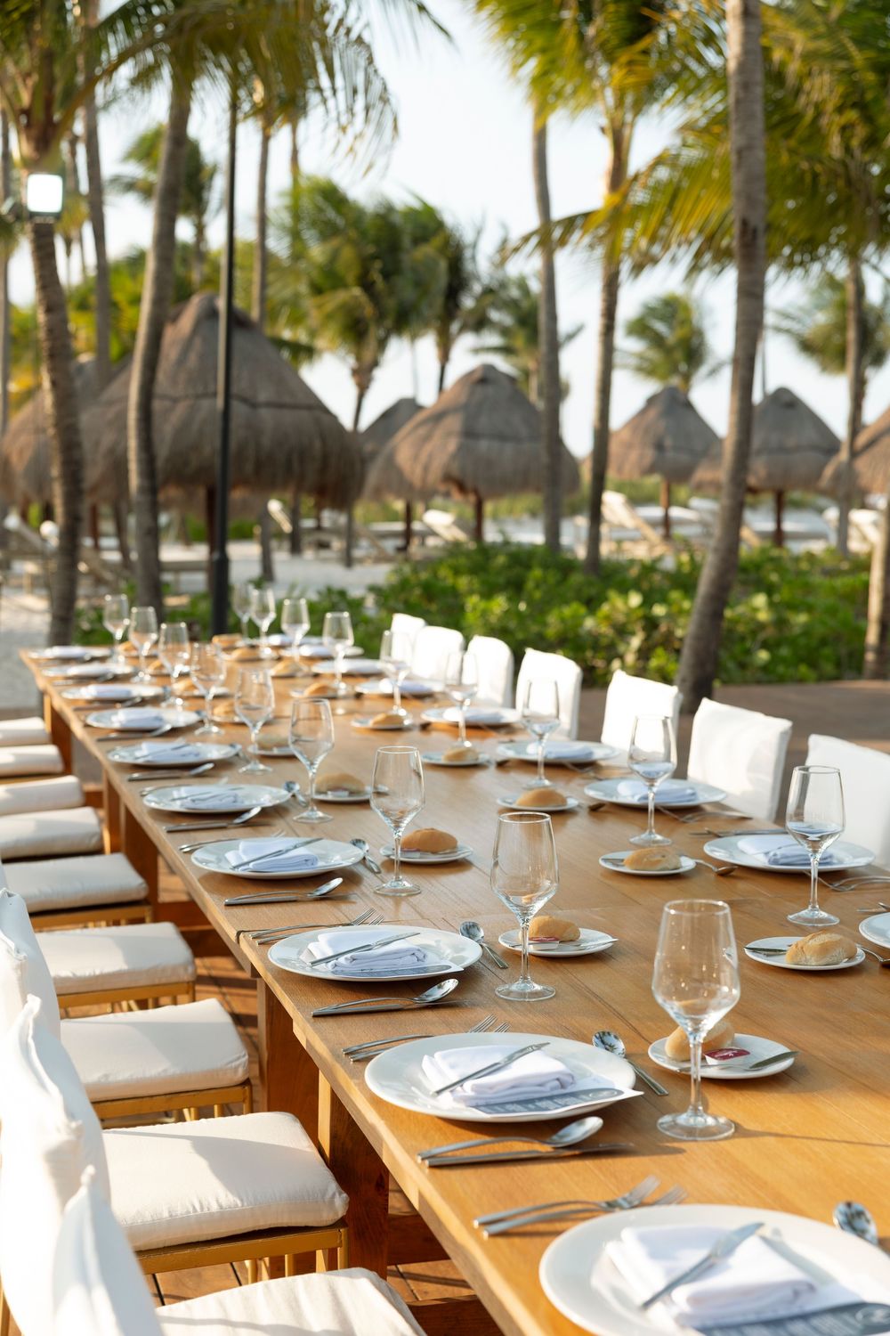 Elegant outdoor dining setup with wooden tables and white place settings under palm trees at a tropical resort.