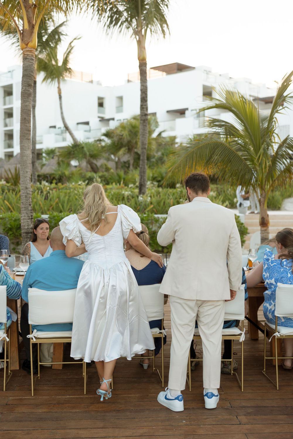 Wedding guests seated at outdoor reception tables with palm trees and white buildings in the tropical background.