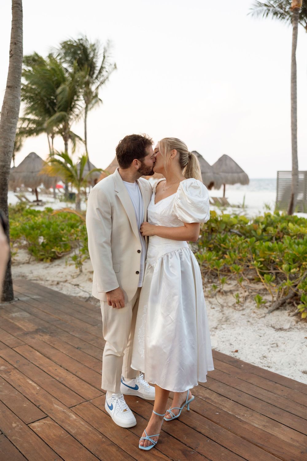 A couple in white and beige outfits share a romantic moment on a beachside boardwalk.