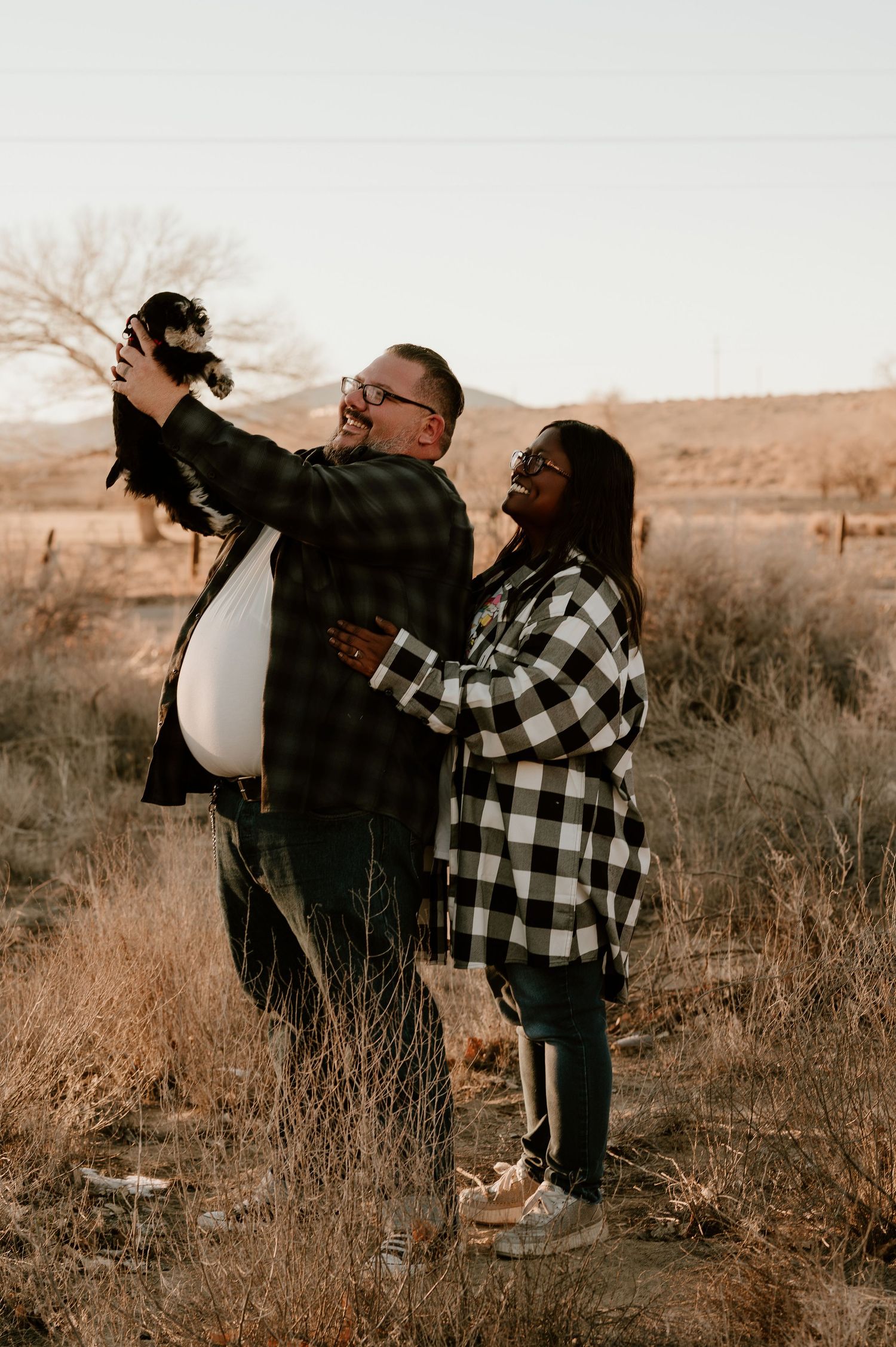 A couple in buffalo plaid embraces in a sepia-toned field during sunset, creating a romantic outdoor moment.