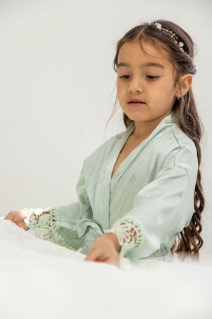 A young person in a mint green robe poses gracefully on white bedding with flowers in their hair.