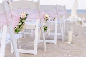 White folding chairs decorated with pink ribbons and greenery line up along the beach for an outdoor wedding ceremony.