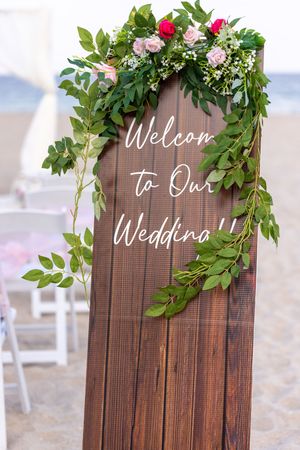 A wooden wedding welcome sign with greenery and floral decorations displayed outdoors with elegant white lettering.