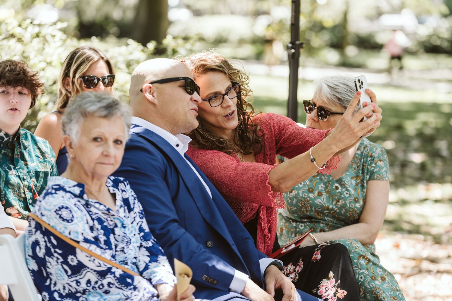A group of people sitting outdoors on white chairs during a sunny garden ceremony.