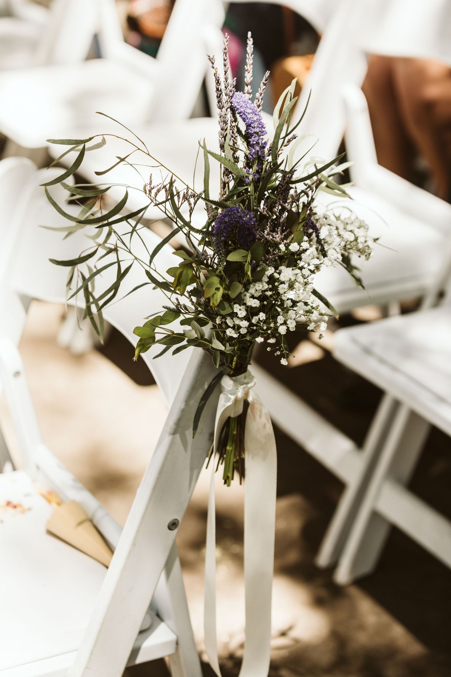 Delicate purple and white floral arrangements adorn white chairs along an outdoor wedding aisle.