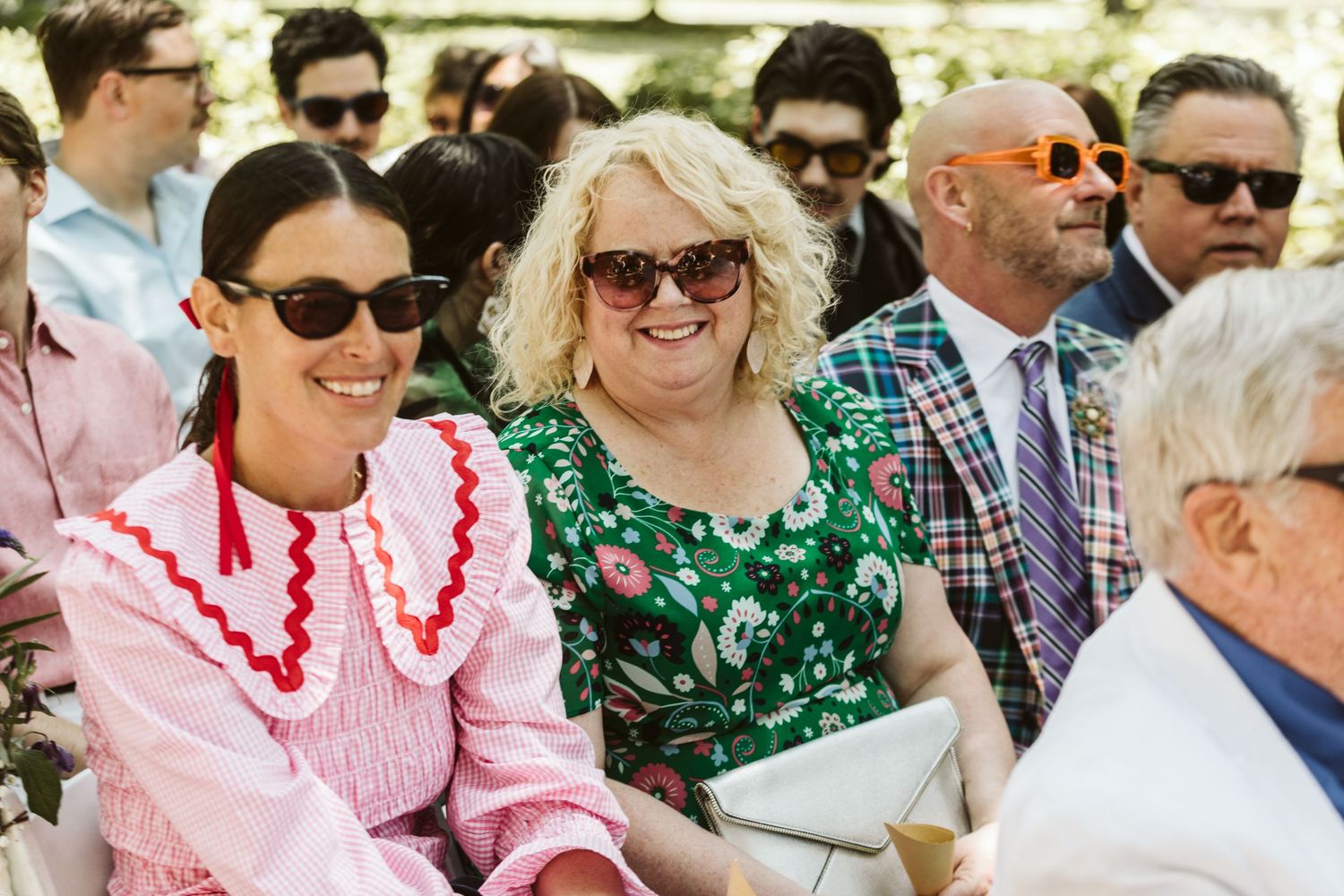 Wedding guests wearing sunglasses and colorful attire smile and chat during an outdoor celebration.