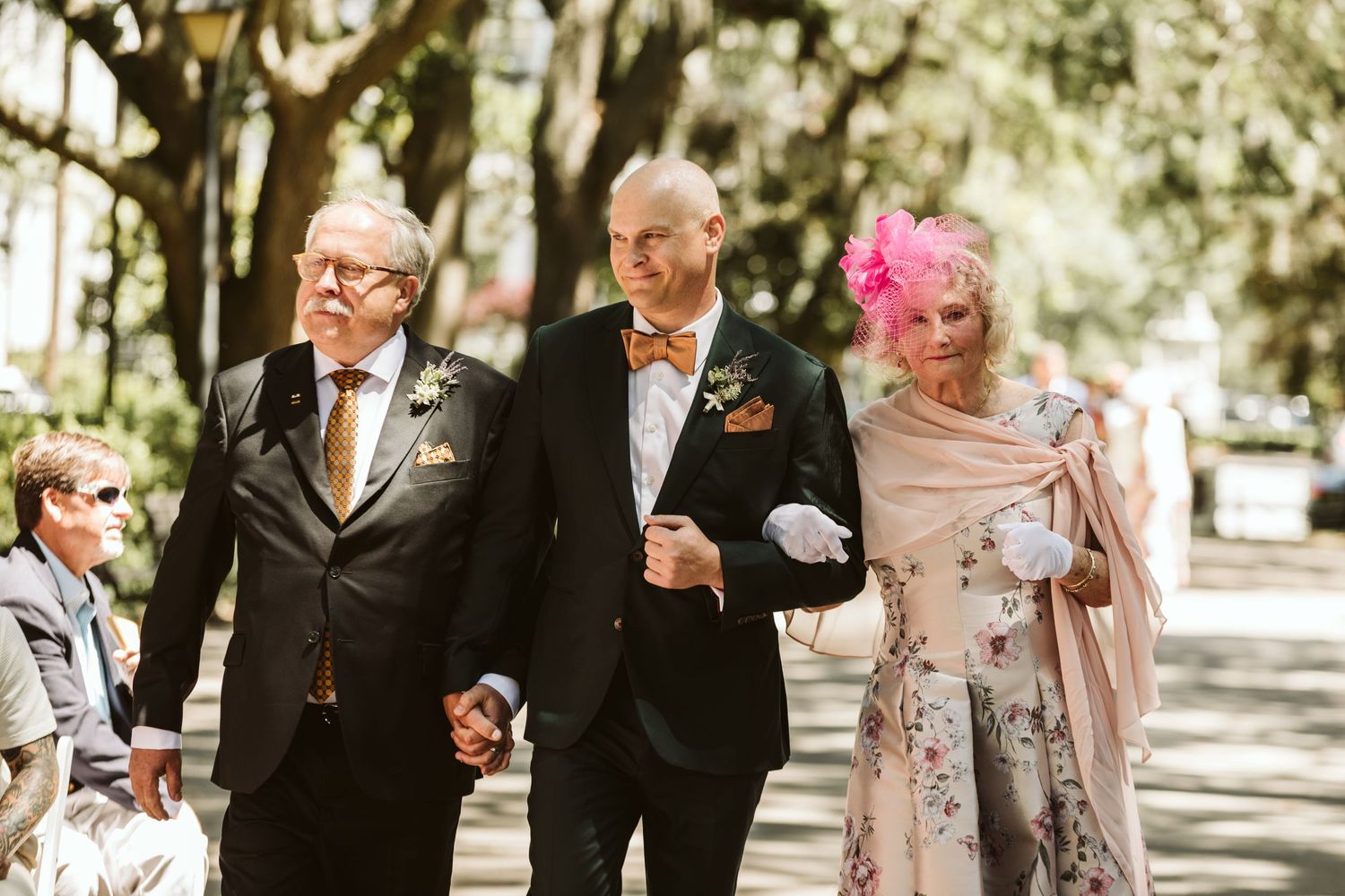 Wedding party members in formal black suits and dresses walk together during an outdoor celebration.
