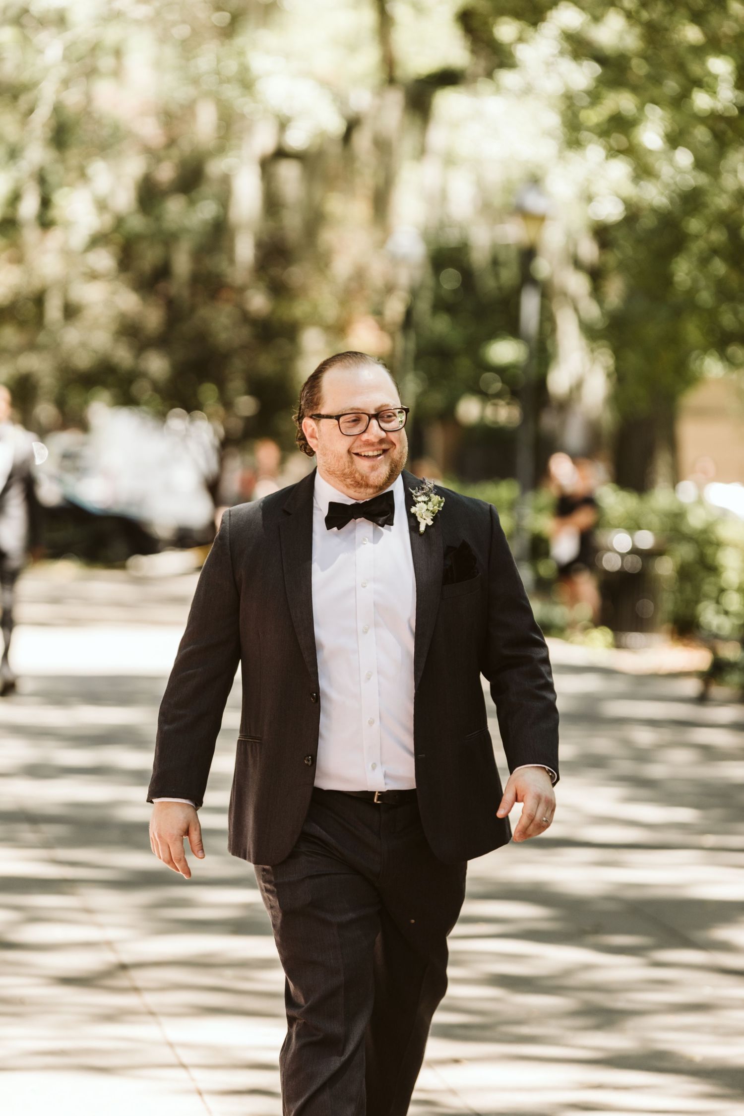 Person in black tuxedo and bow tie walking outdoors on tree-lined path in sunlight.