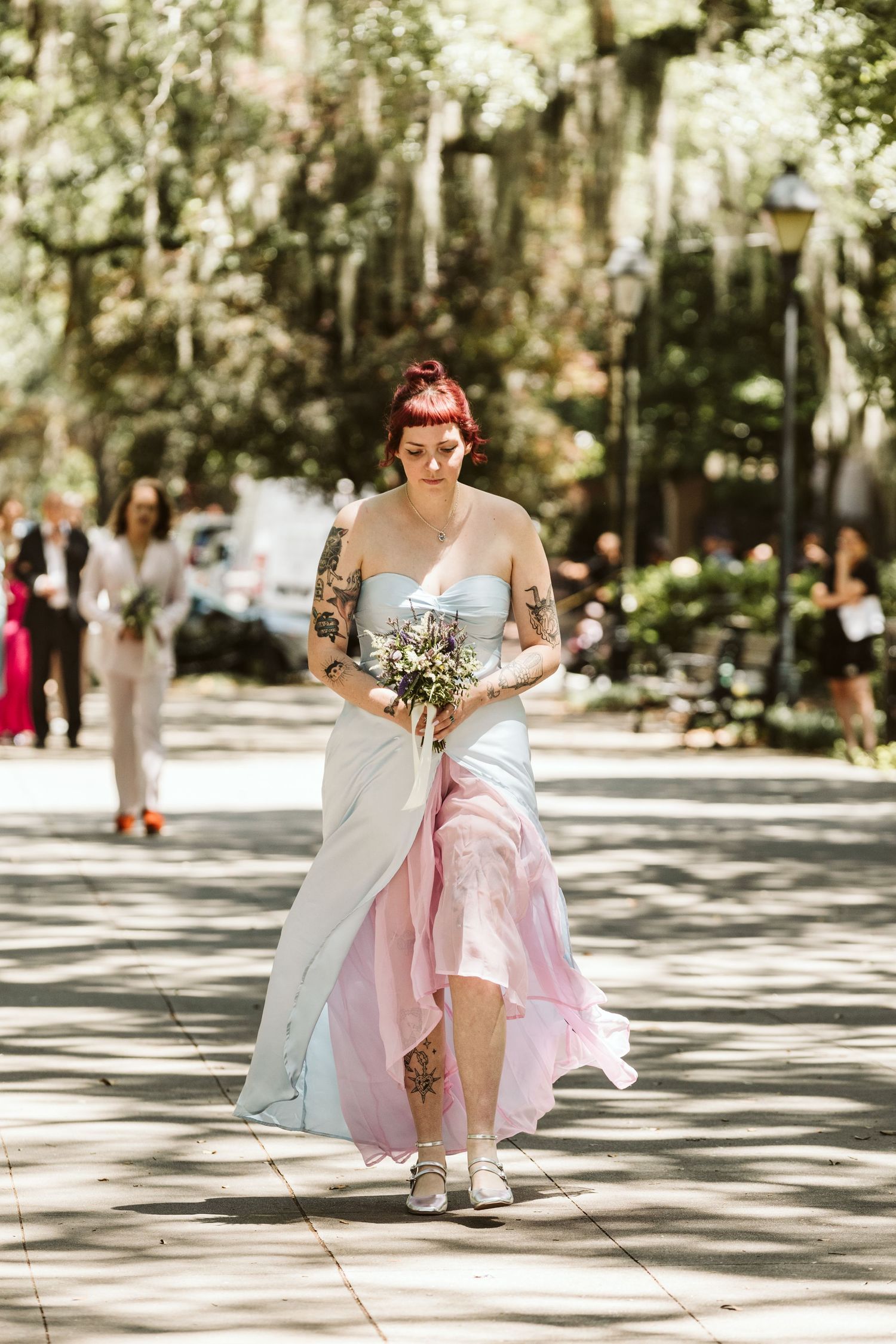 A wedding party member in a pink and white dress sits gracefully on a sunlit path surrounded by trees.