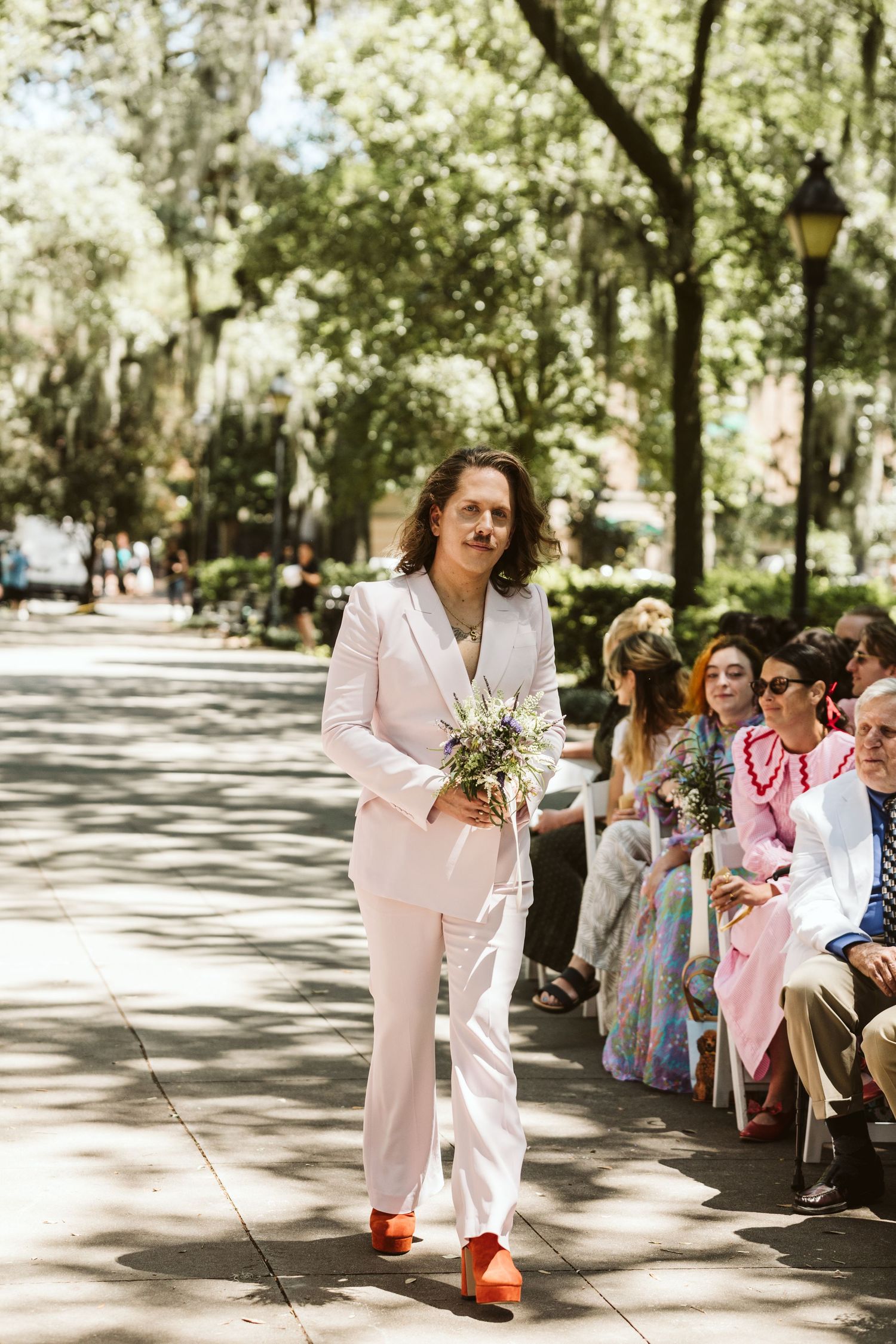 A person in a light pink suit walks down a tree-lined path carrying a small bouquet during an outdoor ceremony.