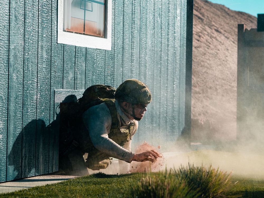 Tactical operator in protective gear crouching by building wall in dusty conditions.