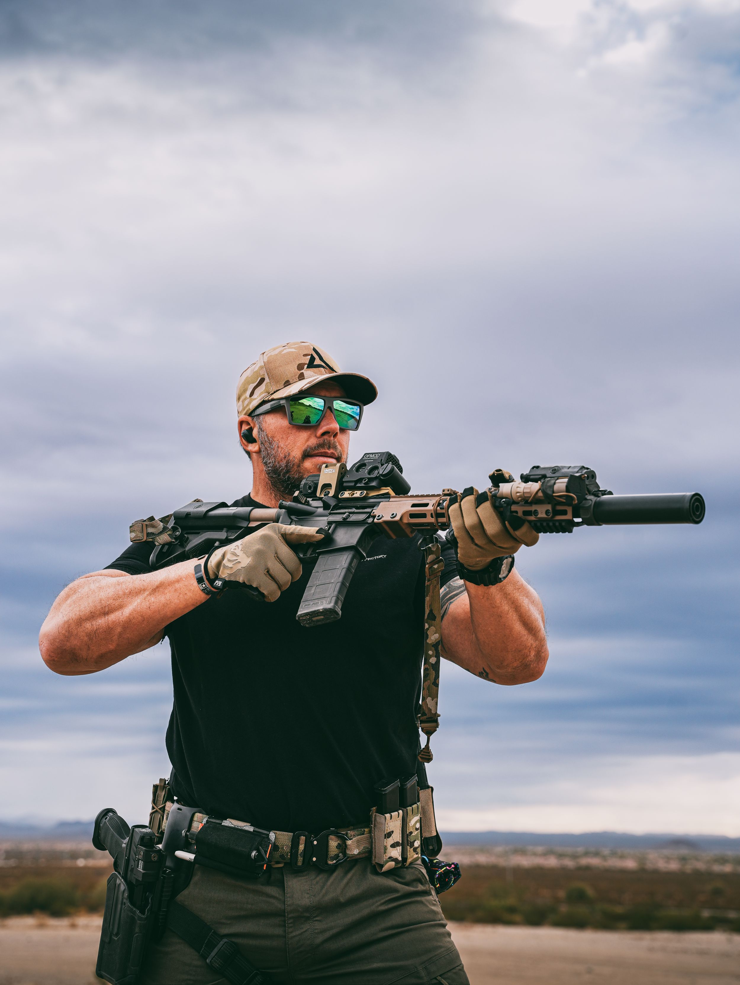 Tactical shooter in black vest aims suppressed rifle at outdoor desert range under cloudy sky.