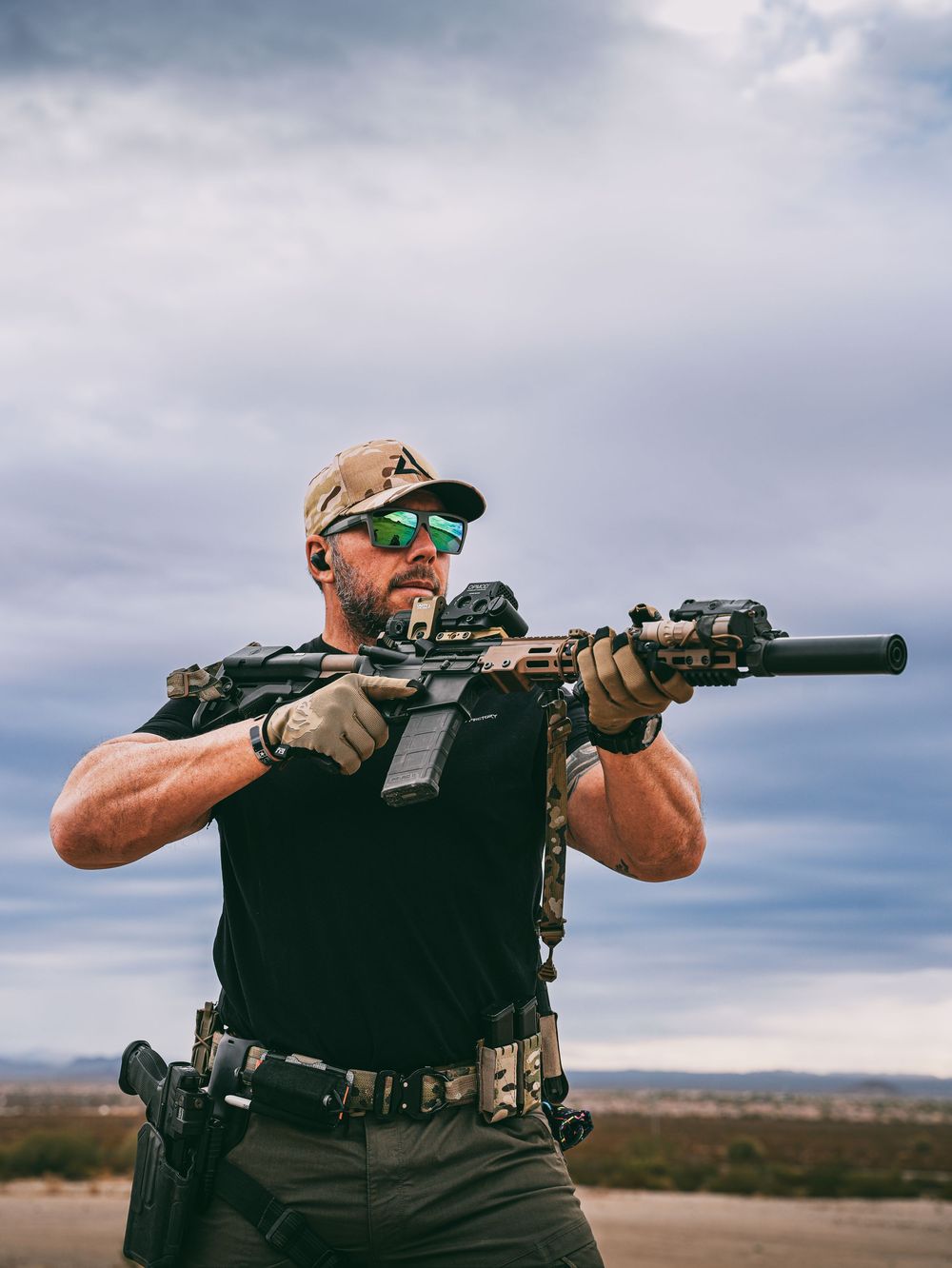 Tactical shooter in black vest aims suppressed rifle at outdoor desert range under cloudy sky.