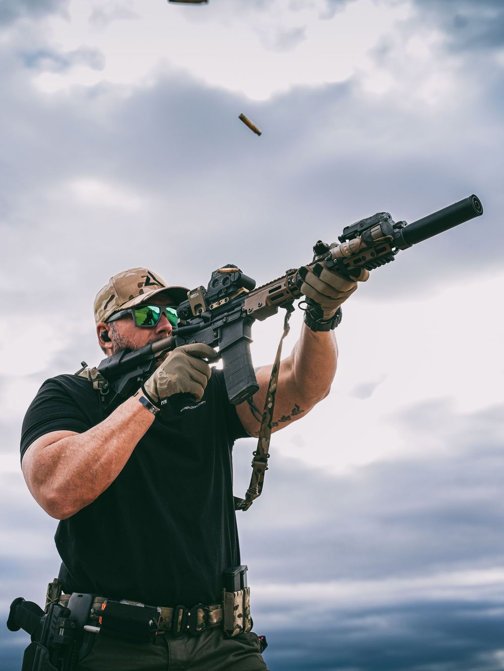 A tactical shooter in black clothing aims an AR-15 style rifle with scope and silencer against a cloudy sky.